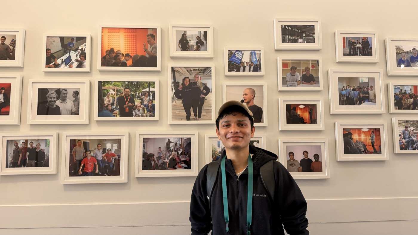 Jaideep in front of a wall of framed photos from YC's history